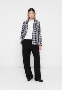 Jonge vrouw met lang haar in een wit shirt, een marineblauwe geruite blouse, zwarte wijde broek en zwarte schoenen, staand tegen een witte achtergrond.
