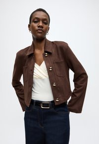 Braune Wildlederjacke mit goldenen Knöpfen, zwei Taschen vorne und einem kurzen Schnitt. Getragen über einem weißen Top und dunkelblauen Jeans.