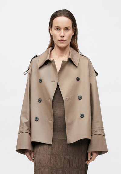 Beige dubbelgeknoopte trenchcoat met een brede kraag, grote zwarte knopen en opgerolde mouwen. Gladde textuur en gestructureerd ontwerp.