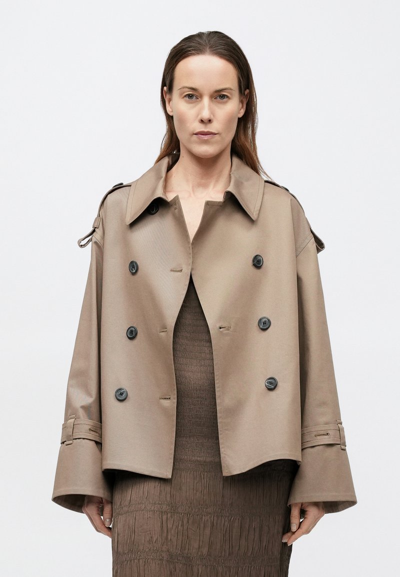 Trench-coat beige croisé à double boutonnage avec un large col, de gros boutons noirs et des manches retroussées. Texture lisse et design structuré.