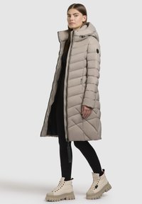 Bežna dolga puffer jakna s kapuco, diagonalni pikasti vzorec, zadrga in stranske žepke, v kombinaciji z bež škornji z debelim podplatom.