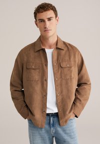 Bruine suède overhemdjas met knoopsluiting en twee borstzakken, gecombineerd met een wit T-shirt en lichtblauwe spijkerbroek. Matte textuur.