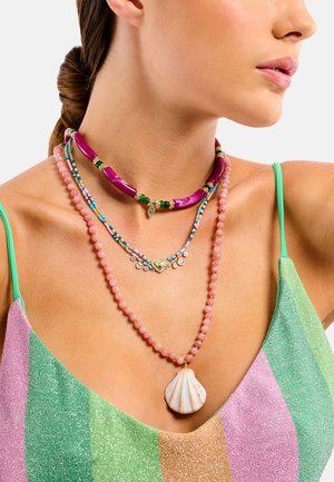 Femme portant trois colliers colorés à plusieurs rangs de perles, dont un avec de grosses perles roses et un pendentif en coquillage, vêtue d'un haut scintillant à rayures.