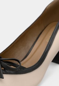 Béžové kožené dámske topánky na nízkom opätku s čiernym lemovaním a malou čiernou mašľou blízko oblého špičky, zobrazené zväčšenie.