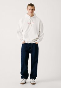 Sudadera blanca con el texto "RED BULL RACING" en rojo, con bolsillo canguro, combinada con pantalones vaqueros azul oscuro y zapatillas blancas con detalles negros.