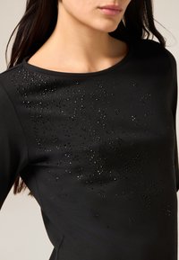 Sort langærmet top med rund halsudskæring og spredte sorte pyntedetaljer foran, lavet af glat, elastisk stof.