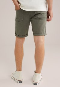 Mies, joka on pukeutunut ylös käärittyihin oliivinvihreisiin shortseihin, valkoisiin korkeaankartioisiin Converse-kengiin ja valkoiseen paitaan, seisoo yksinkertaista taustaa vasten.