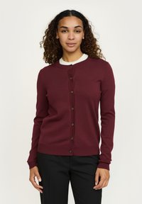 Frau mit lockigem Haar, die eine burgunderrote, geknöpfte Strickjacke über einem weißen Hemd und schwarzen Hosen trägt, steht vor einem schlichten hellen Hintergrund.