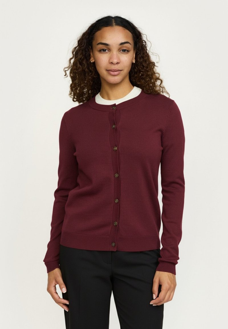 Frau mit lockigem Haar, die eine burgunderrote, geknöpfte Strickjacke über einem weißen Hemd und schwarzen Hosen trägt, steht vor einem schlichten hellen Hintergrund.