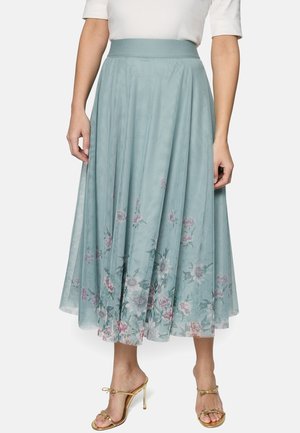 Femme portant une jupe midi bleu clair avec un ourlet à imprimé floral, associée à des sandales à talons hauts dorées et un haut blanc.