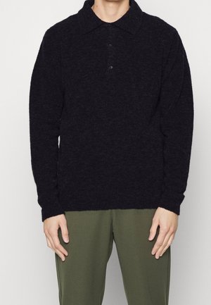 Muž v černém polo svetru s dlouhým rukávem a olivově zelených kalhotách, stojící s rukama volně podél těla.