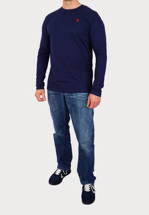 Chemise à manches longues bleu marine avec un logo rouge, associée à un jean bleu et des baskets bleu marine. Coupe confortable, texture lisse et style décontracté.