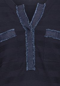 Gros plan d'un tissu tricoté bleu foncé avec des rayures texturées et une bordure en denim effilochée le long d’un col en V et deux accents horizontaux sur la poitrine.