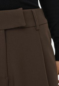 Braune Hose mit einem breiten Bund und gesteppten Details. Das Material wirkt glatt mit einem mittleren Glanz. Sie hat Falten an der Vorderseite.