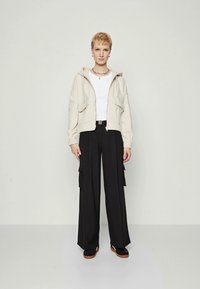 Person med kort blondt hår iført en lys beige hettegenser, hvit t-skjorte, svarte vide cargobukser og svarte sko med brune såler.