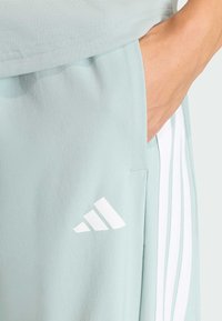 Pantalon de sport vert clair avec des rayures blanches et un logo Adidas. Présente des poches latérales et une texture douce et lisse.