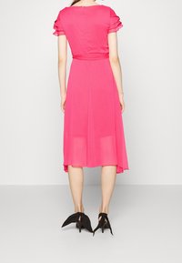 Robe rose fuchsia avec manches courtes à volants et corsage ajusté. La jupe fluide arrive au milieu du mollet, avec une légère brillance. Talons noirs avec lacets.