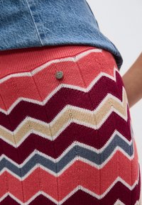 Κοντινό πλάνο πολυχρωμικής πλεκτής φούστας με ζικ-ζακ σχέδιο σε κοράλλι, μπορντό, μπεζ και γκρι, σε συνδυασμό με μπλε τζιν τοπ.