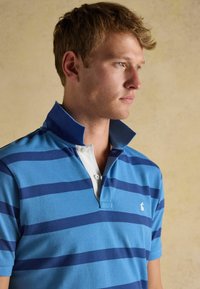 Blaue Poloshirt mit horizontalen marineblauen Streifen und weißer Knopfleiste. Hat einen hohen Kragen und ein gesticktes Logo auf der Brust. Aus Baumwollmaterial.