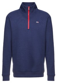 Marinblå fleece-pullover med hög krage, featuring en röd halvdragkedja och en liten logo på vänster br öst. Ribbad mudd och fåll.