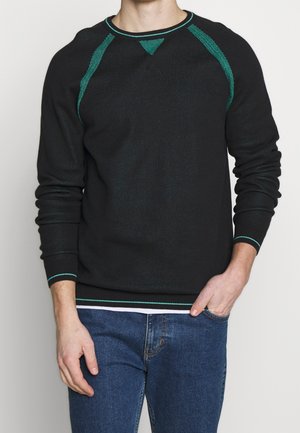 Homme portant un pull à manches longues noir avec des bordures vertes, un jean bleu, une main dans la poche, devant un fond blanc uni.