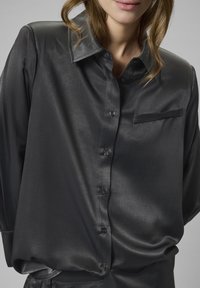 Kvinde iført en sort satin skjorte med knapper og krave samt brystlomme, vist fra skuldrene til hoften mod en neutral baggrund.