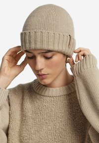 Beige stickad mössa med ribbad mudd, gjord av texturerad ull, bärs av en person. Matchande beige tröja med rund halsringning och lös passform.