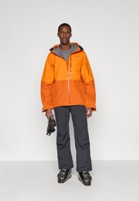 Mies, joka käyttää oranssia ja harmaata hiihtotakkia sekä -housuja, pitää hiihtogogglesia kädessään seistäkseen hiihtokengissä yksinkertaista taustaa vasten.