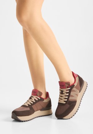 Benen met bruine en beige sneakers met rode accenten en crèmekleurige veters, staand op een effen lichte achtergrond.