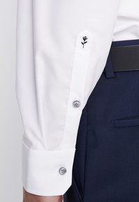 Hvid langærmet skjortemanchet med to hvide knapper og lille sort broderet rosedetalje, kombineret med mørkeblå bukser og sort bælte.