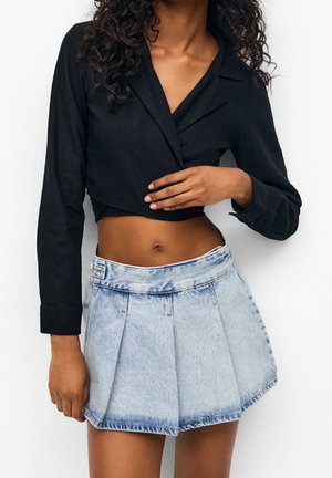 Vrouw die een zwarte cropped top met lange mouwen en een lichtblauwe hooggetailleerde geplooide denim mini-rok draagt, staand tegen een witte achtergrond.