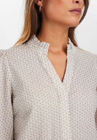 Balta blusa su mažais violetiniais gėlių raštais, su mandarinų apykakle ir raukšlėta iškirpte. Pagaminta iš lengvos medžiagos su lygia tekstūra.
