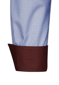 Tessuto blu a motivi con piccoli triangoli e un polsino marrone. Il materiale appare liscio e leggero, mostrando una texture e un design distintivi.