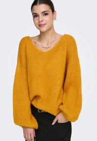 Oversized strikkegenser i sennepsgul med dyp V-hals, teksturert stoff og ballongermer, kombinert med svarte bukser og minimalistisk smykke.