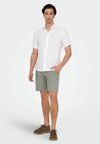 Chemise blanche à manches courtes avec un col, assortie à un short gris. Modèle portant des chaussures en daim marron, mettant en avant une tenue décontractée pour l'été.