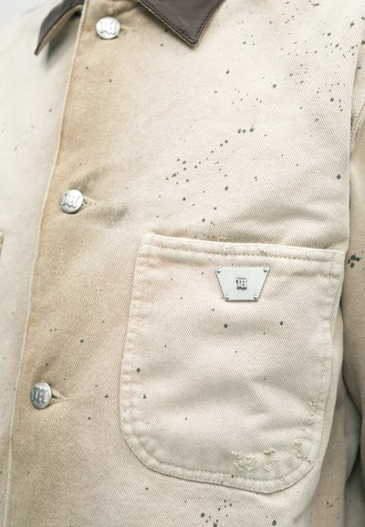Chemise en toile beige avec col marron, boutons argentés et une plaque logo triangulaire sur la poche. Présente des taches sombres éparpillées et une finition texturée.