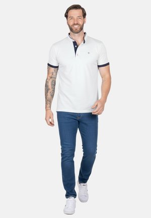 Hombre con barba y tatuaje caminando hacia adelante, vistiendo una camiseta polo blanca, jeans azules y zapatillas blancas, sobre un fondo blanco liso.