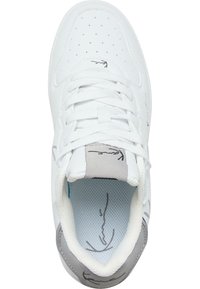 Hvit sneaker med perforert tåboks, grå detaljer på hælen, flate lisser, og et trykt design på tungen. Teksturert innersåle synlig.