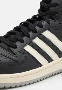 Czarne skórzane sneakersy z trzema białymi teksturowanymi paskami, płaskimi czarnymi sznurowadłami i gumową podeszwą z delikatnym szarym wzorem bieżnika.