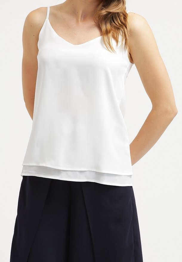 Femme portant un haut blanc sans manches à col en V associé à un pantalon plissé noir taille haute sur un fond uni.