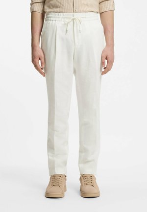 Hombre vistiendo pantalones blancos con cordón, camisa de rayas beige y zapatillas beige con cordones, de pie contra un fondo blanco.