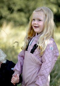 Šviesiai rožinė dygsniuota striukė su aukštu apykakliu ir juodomis dirželiais, derinama su atitinkančia šviesiai rožine kombinezonu. Ilgi šviesūs plaukai ir laikosi už rankų.