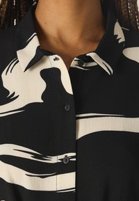 Camicia a maniche lunghe bianca e nera con un motivo astratto a onda. Tessuto testurizzato con chiusura a bottoni e una leggera lucentezza.