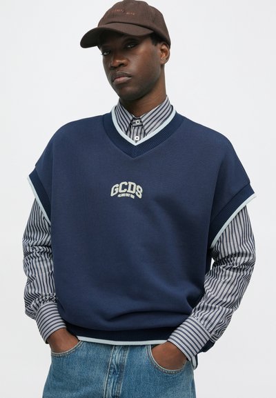 Sweater oversized azul-marinho com decote em V, logótipo bordado "GCDS", combinado com uma camisa riscada e jeans de ganga, enfeitado com um boné castanho.