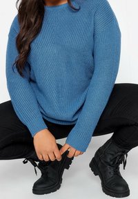 Blå stickad tröja med ribbade manschetter, tillsammans med svarta snörstövlar. Tröjan har en lös passform och en texturerad mönster.