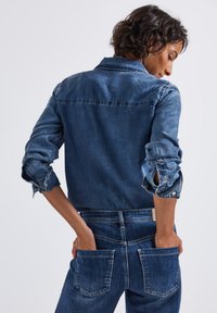 Dunkelblaue Jeanshemd mit spitzem Kragen, hochgekrempelten Ärmeln und kontrastierenden Nähten. Kombiniert mit passenden Jeanshosen mit Gesäßtaschen.