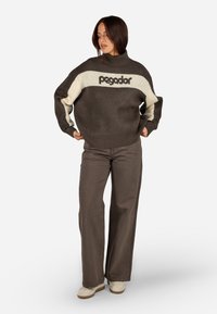 Rudas megztinis su kreminiu akcentu ir logotipu; golfo stiliaus; plačiomis rankovėmis; derinamas su taupe spalvos plačiomis kelnių ir bežiniais sportbačiais.