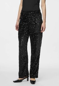 Pantalons en sequins noirs à coupe droite et taille élastique, avec une surface brillante et texturée. Associés à des chaussures à talons noires.