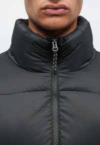 Μαύρο μπουφάν με ψηλό γιακά, με μπροστινό φερμουάρ και λεία, γυαλιστερή υφή. Μαλακή υφή με καπιτονέ τμήματα.