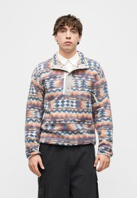 Νεαρός άνδρας που φοράει πολύχρωμο φλις πουλόβερ με λευκό πουκάμισο και μαύρο παντελόνι cargo, στέκεται μπροστά σε ένα απλό φόντο.